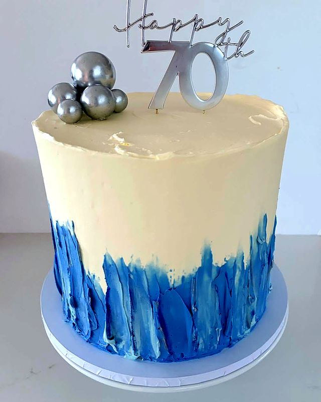 A tall cream buttercream cake with blue palette-knife strokes around the base, silver pearls and a silver Happy 70th topper.