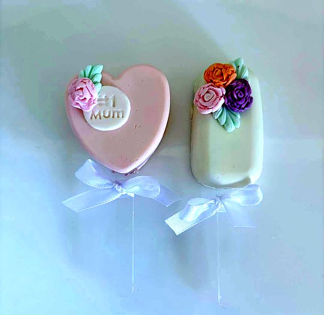 Two cakesicles with white ribbons, a pink heart with a #1 Mum plaque and a white bar with piped roses.