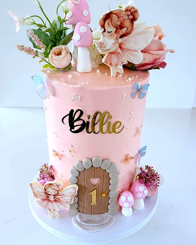 A tall pink buttercream cake with a little wooden door marked 1, butterflies, toadstools, flowers and two fairies.