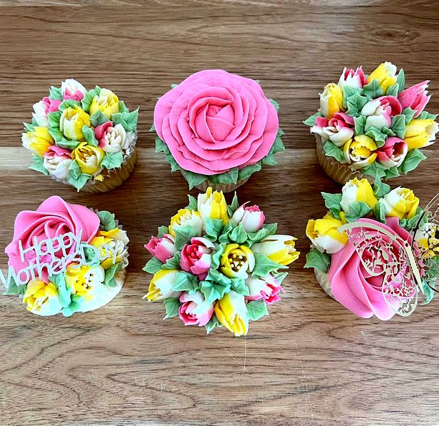 Six cupcakes seen from above on a wooden table, piped with pink roses, tulips and green leaves.