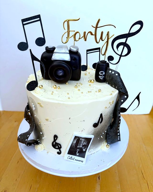 A cream cake with black music notes, a black camera, a film strip and a gold Forty topper.